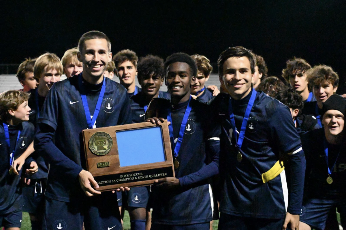 WIN TOGETHER. Team captains Liam Sullivan, Ezra Straub, and Arlo Zirps smile for the camera with their placard. After photos were finished, the team celebrated by hoisting the trophy high into the air in front of the attending student section, who responded with an echoing cheer.