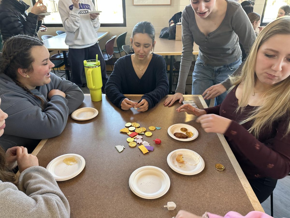 EMERGE VICTORIOUS. Juniors Helen Frost, Paloma Gomez-Whitney, Hazel McCarthy, and Sophie Donahue (left to right) gather in a circle playing dreidel. The students are competing to win the chocolate coins and stickers in the middle of the table. 